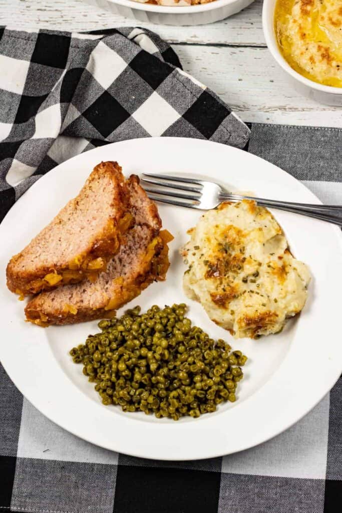 serving of Amish hamloaf on white plate with peas and mashed potatoes